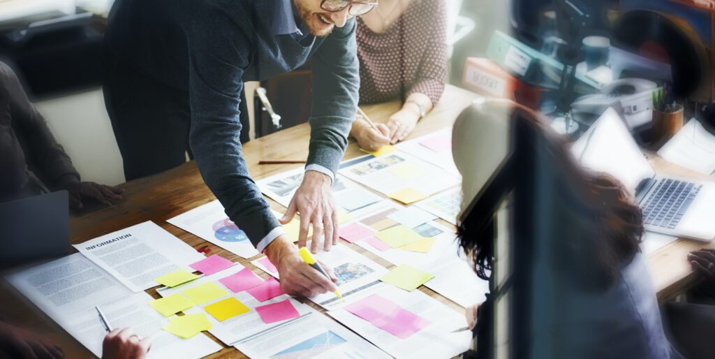 A landscape image of a a group content design session being held. One person stands over a table which has many different pieces of paper. Pink and yellow post it notes are also covering the pieces of paper. We can see the session has two women and three men. In the background, there are two binders, red and green, in colour on a desk. 