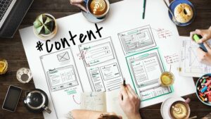 A landscape image of a content design group session discussing content. There are pieces of paper on the table, glasses of drinks and a bowl of sweets to the right. The paper shows three rectangle wireframes of web pages