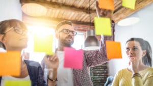 A landscape image of three colleagues working together. Pink, orange and yellow post-it notes are placed on a clear board.