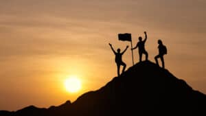 Landscape image of three people atop a mountain. The sun is visible in the background as the three people celebrate completing their mission to reach the top.